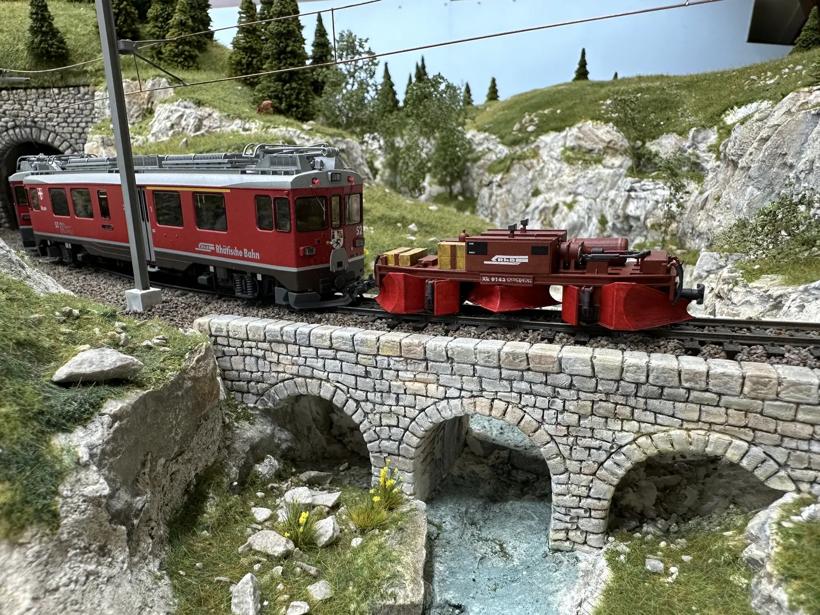 Red locomotive 802 and a plough vehicle crossing a stone model railway bridge